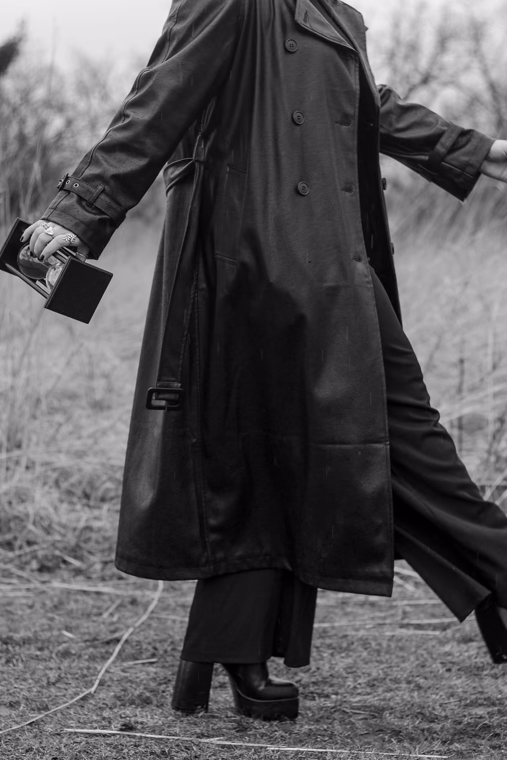 Hannah Hernandez walking in a field in a long black trench coat and tall heels while holding an hourglass.