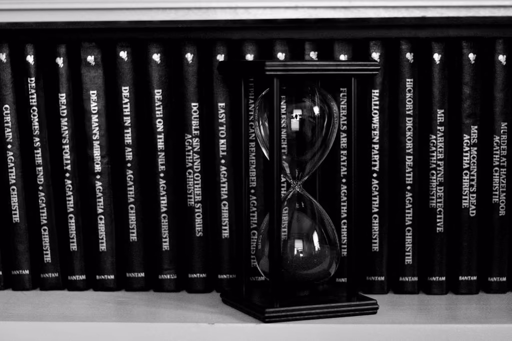 An hourglass sitting on a bookshelf filled with murder mystery books written by Agatha Christie.