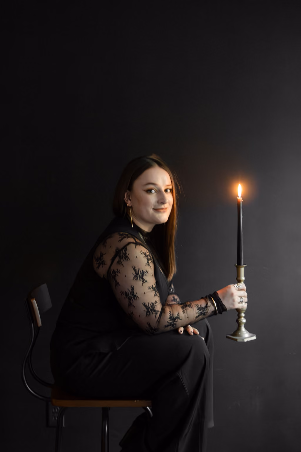 Hannah Hernandez sits on a stool in front of a black wall. She holds a lit candle in her hand.