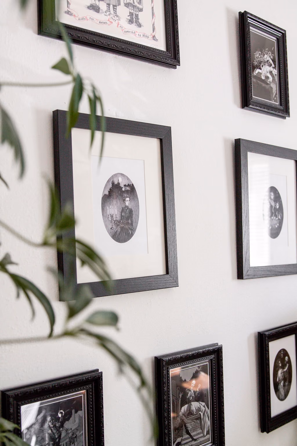 Several pictures hang on a white wall with a small part of a plant poking into the image. The 7 frames portray macabre scenes.