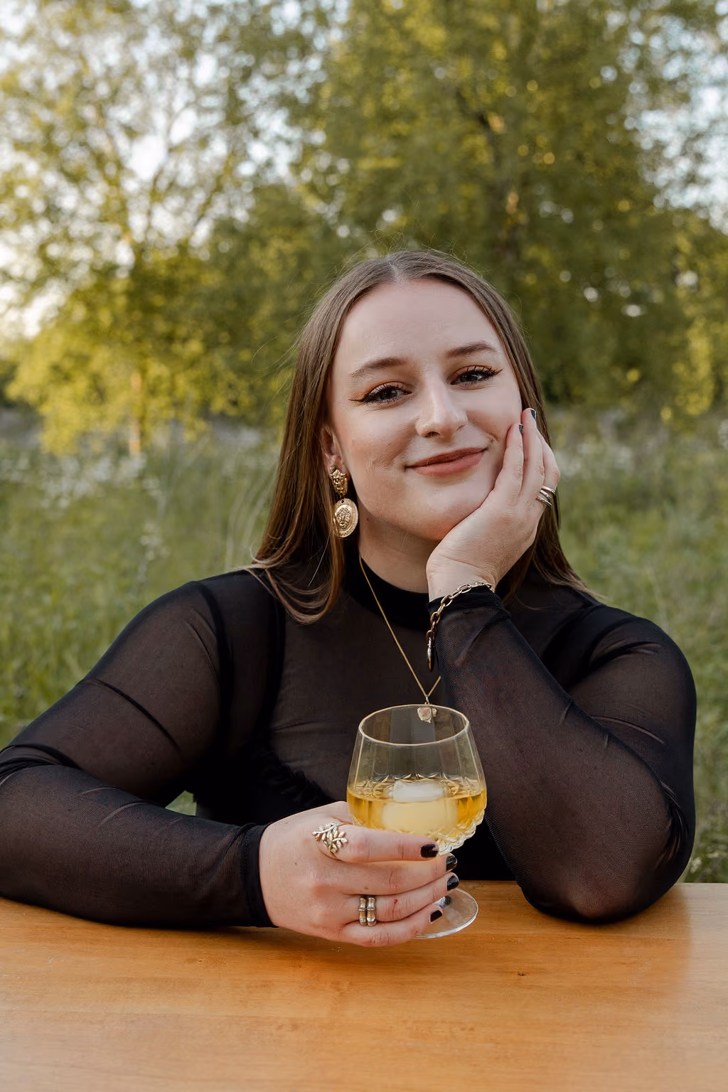 Hannah Hernandez in black sheer top holding a drink with ice while sitting outdoors at a wooden table.