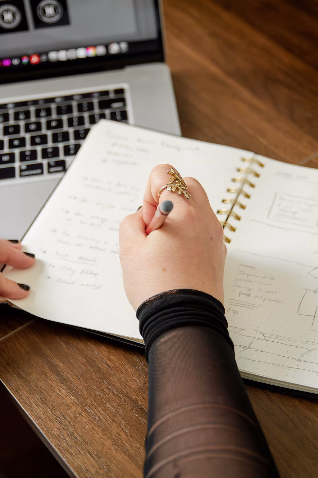 Hand with decorative ring writing notes in a spiral notebook beside a laptop on a wooden table.
