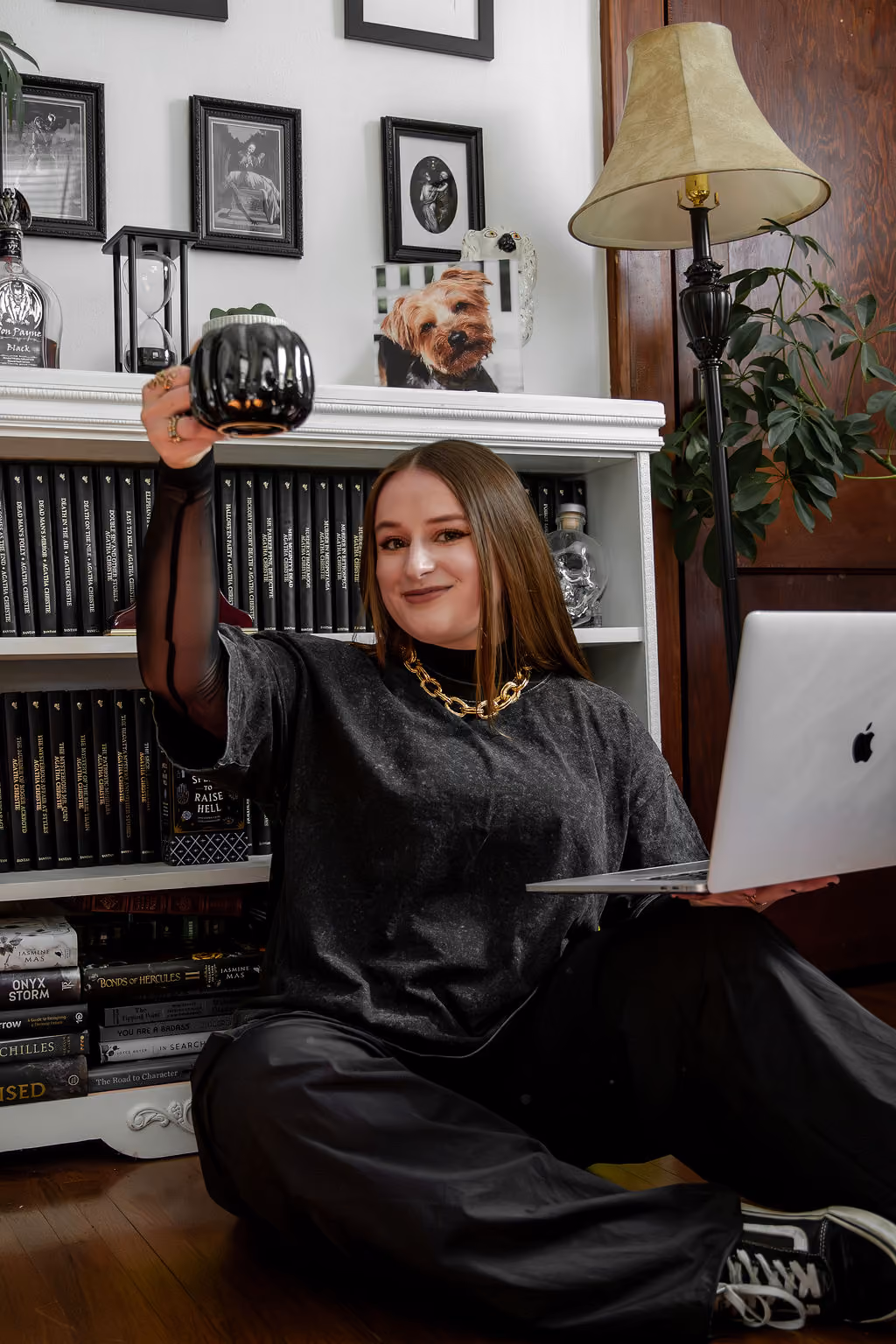 Smiling woman sits on floor holding a black mug and laptop in a cozy room with books and dog photos.
