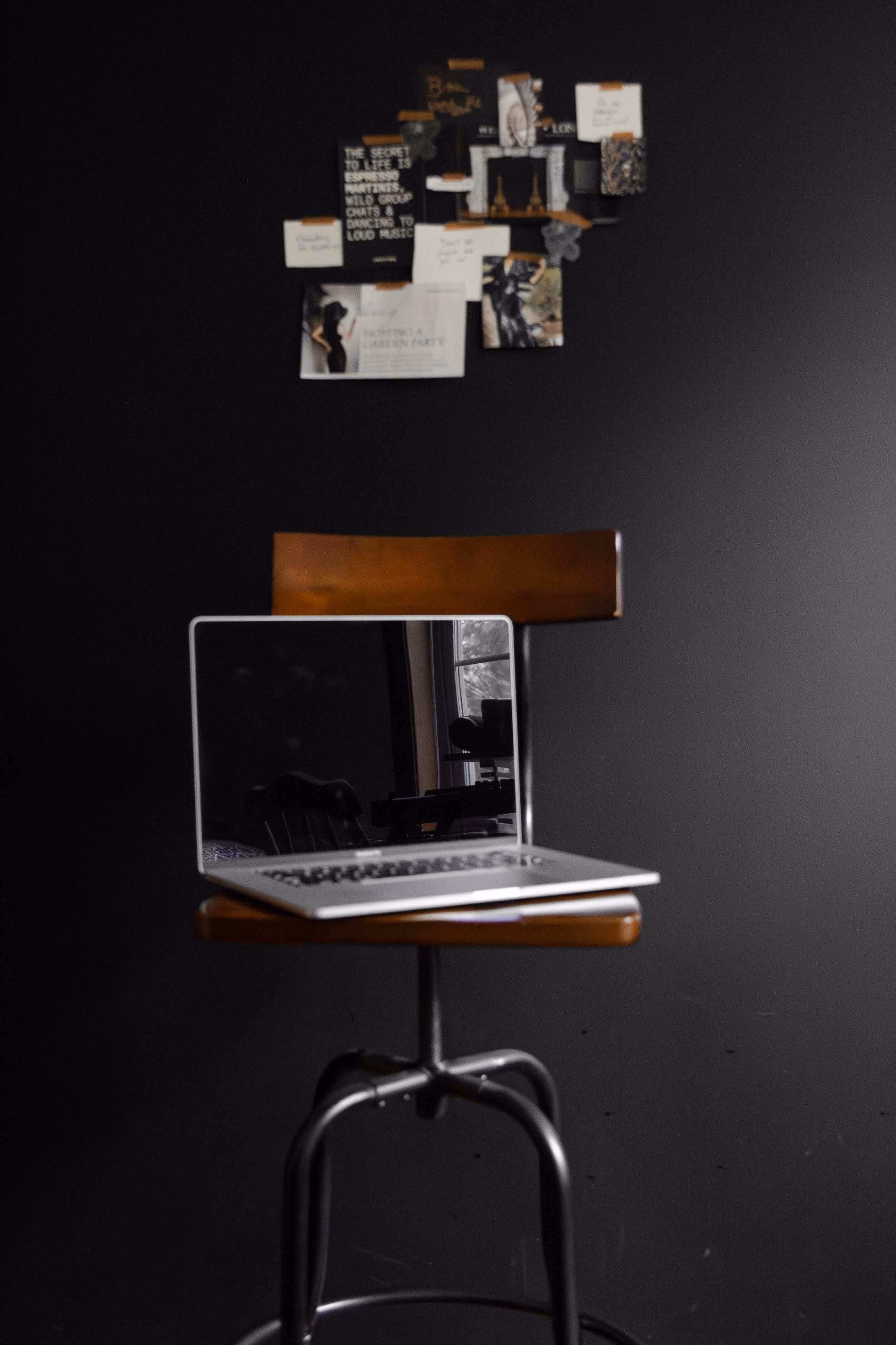 Silver laptop with black screen on wooden chair against a dark wall with pinned notes.