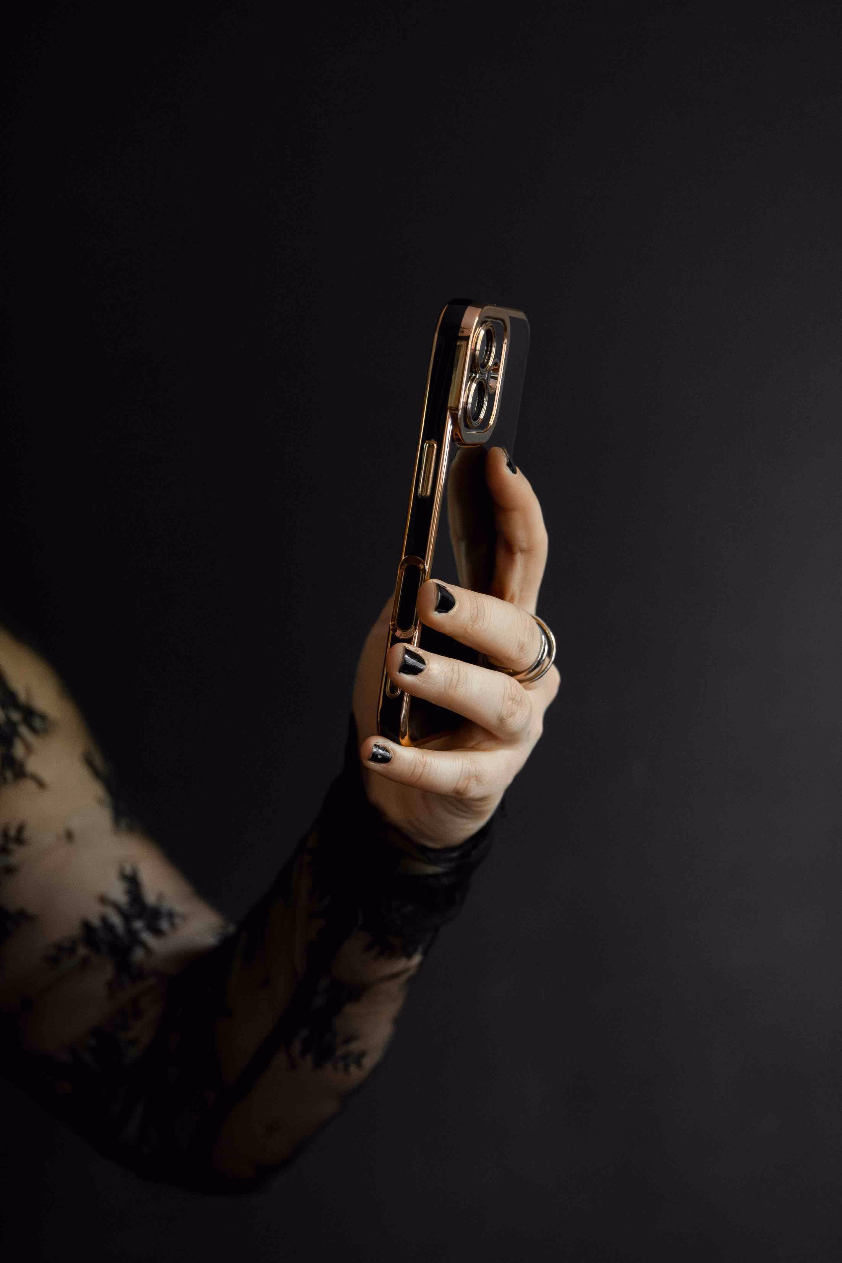 Hand with black nail polish holding a smartphone with a reflective gold case against a dark background.