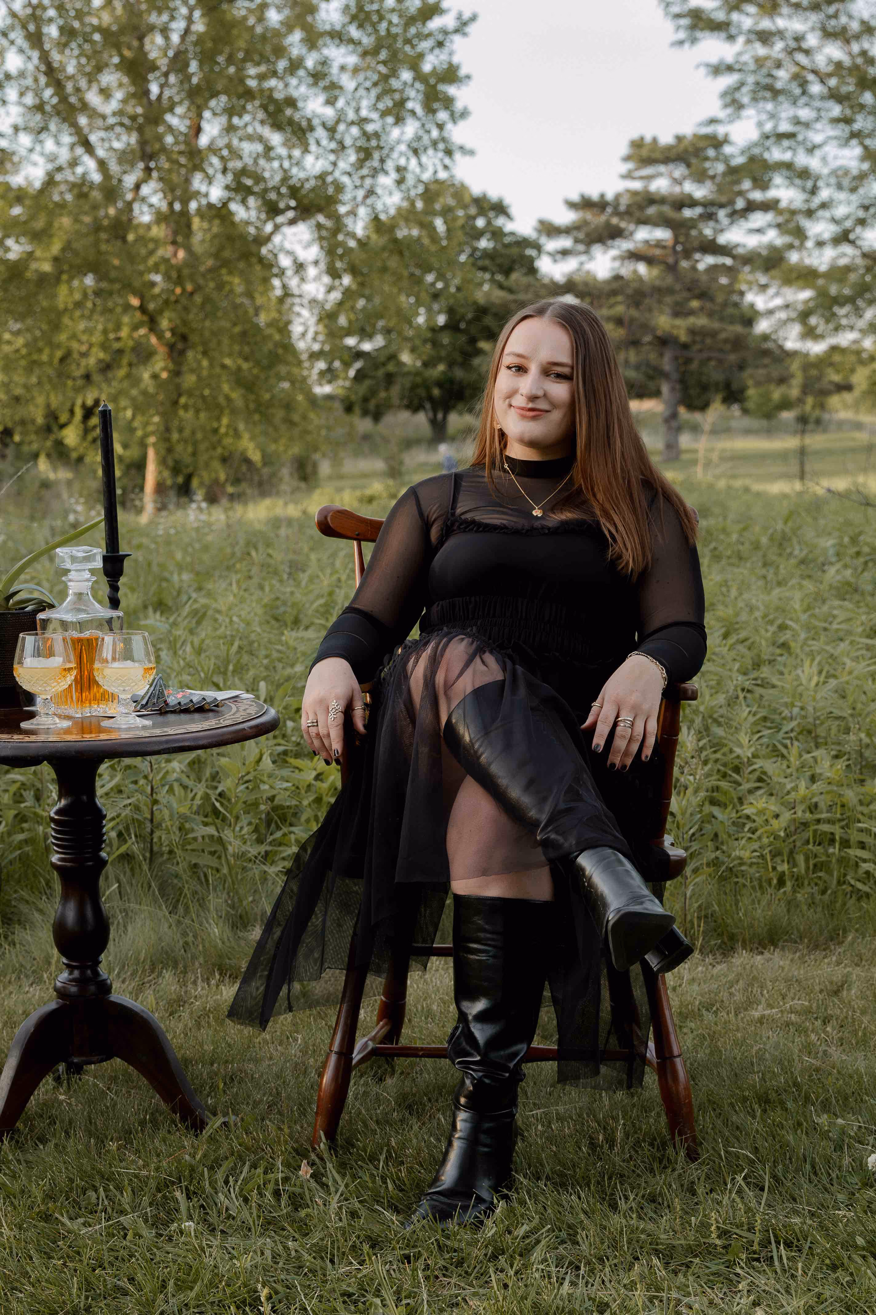 Woman in black dress and boots sitting on wooden chair outdoors near table with drinks and candlestick.