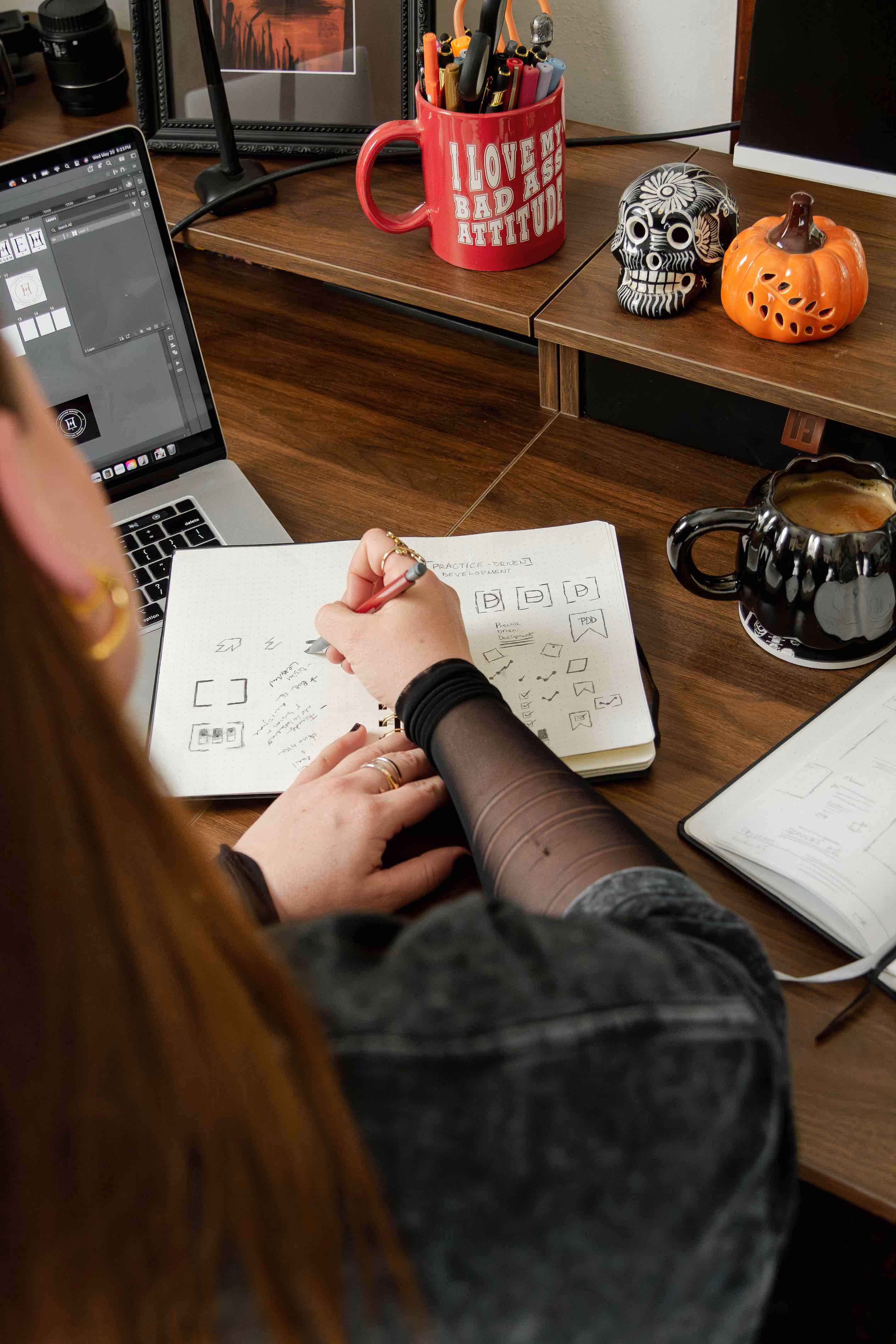 Person writing notes and sketches in a notebook at a wooden desk with a laptop and decorative items.