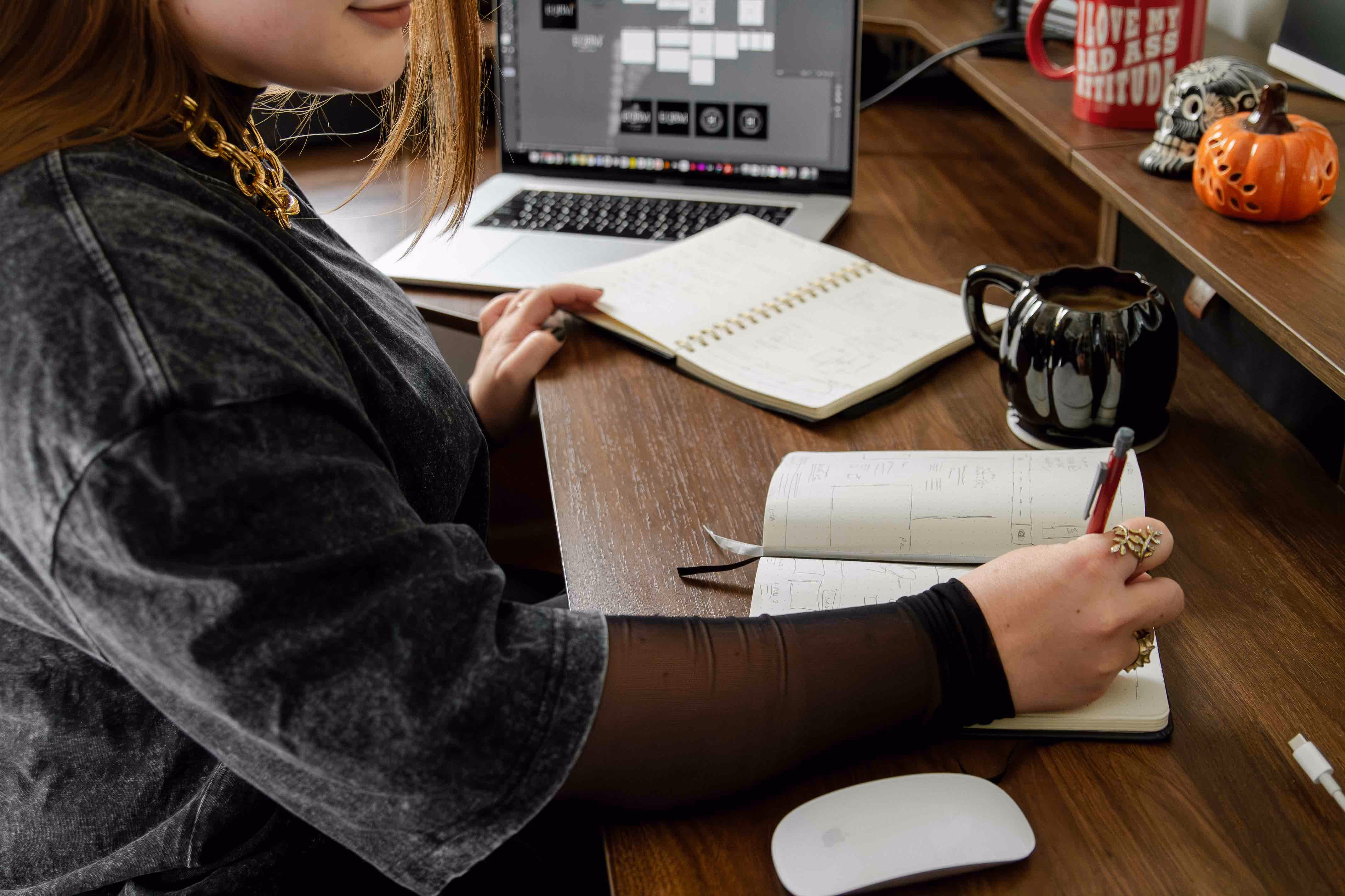Person writing in a notebook at a wooden desk with a laptop, coffee mug, and pumpkin decor.