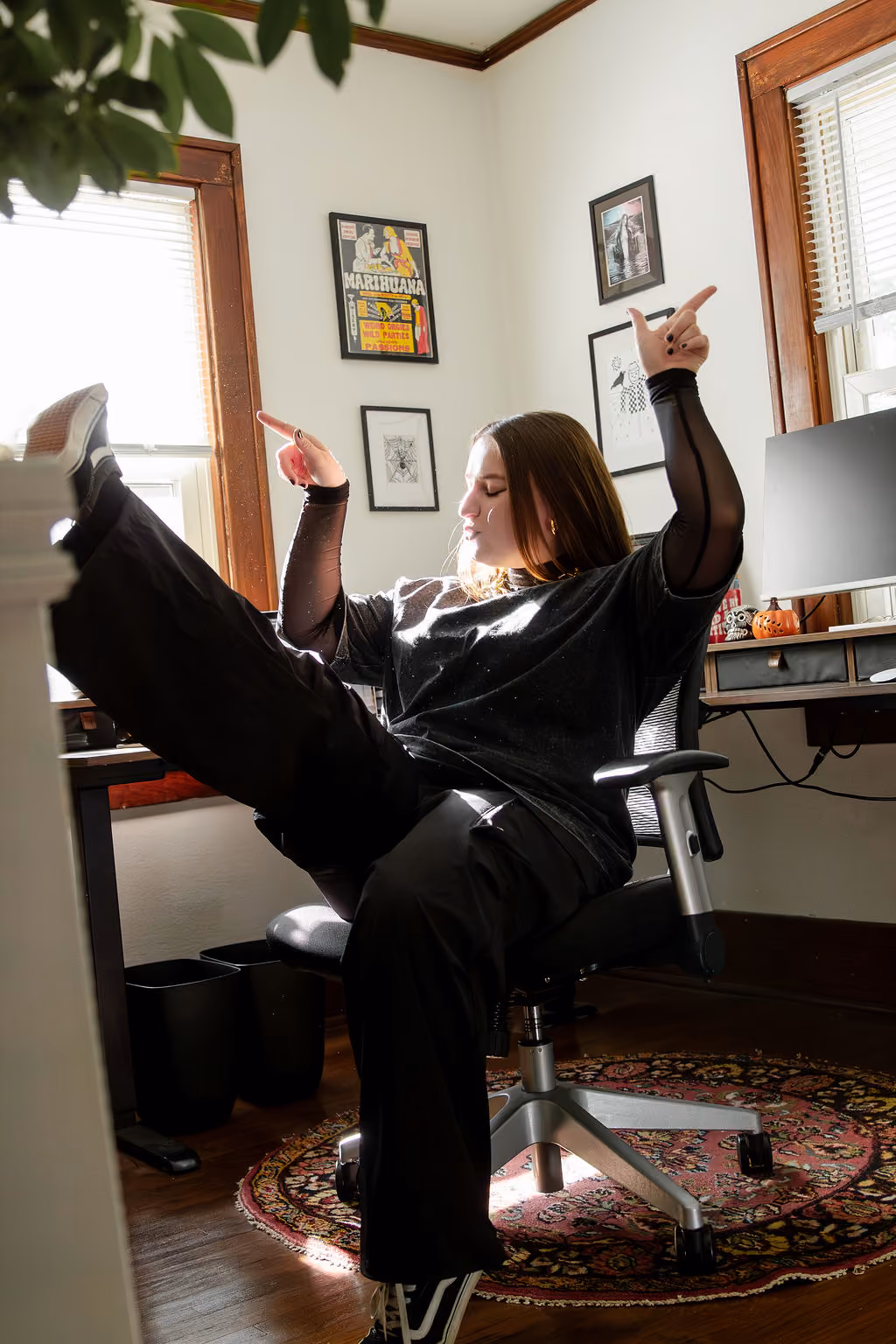 Person with long hair in black clothes sitting on office chair with legs raised and fingers pointing up.
