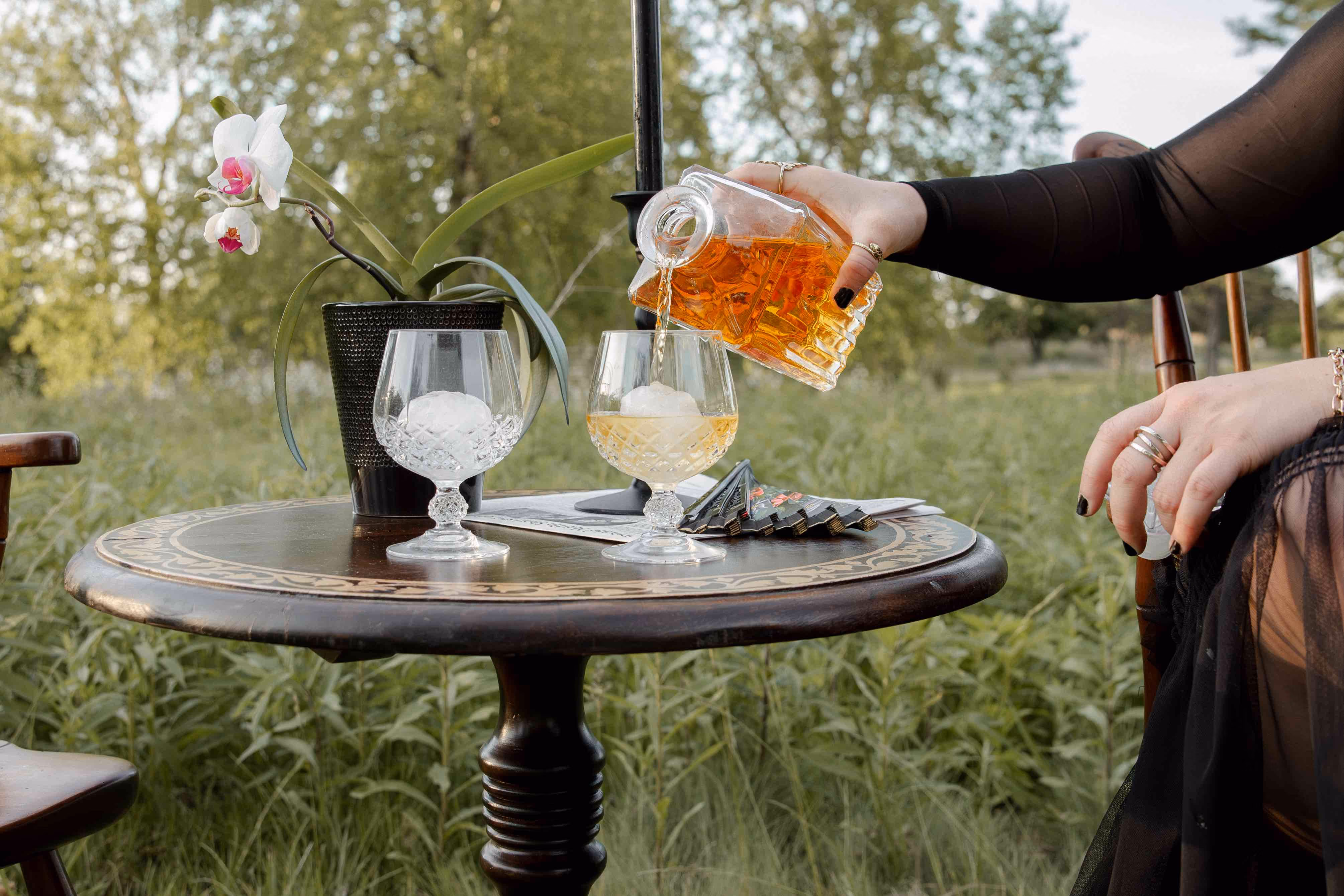Hand pouring amber liquor from a bottle into a glass with ice on a wooden table outdoors.