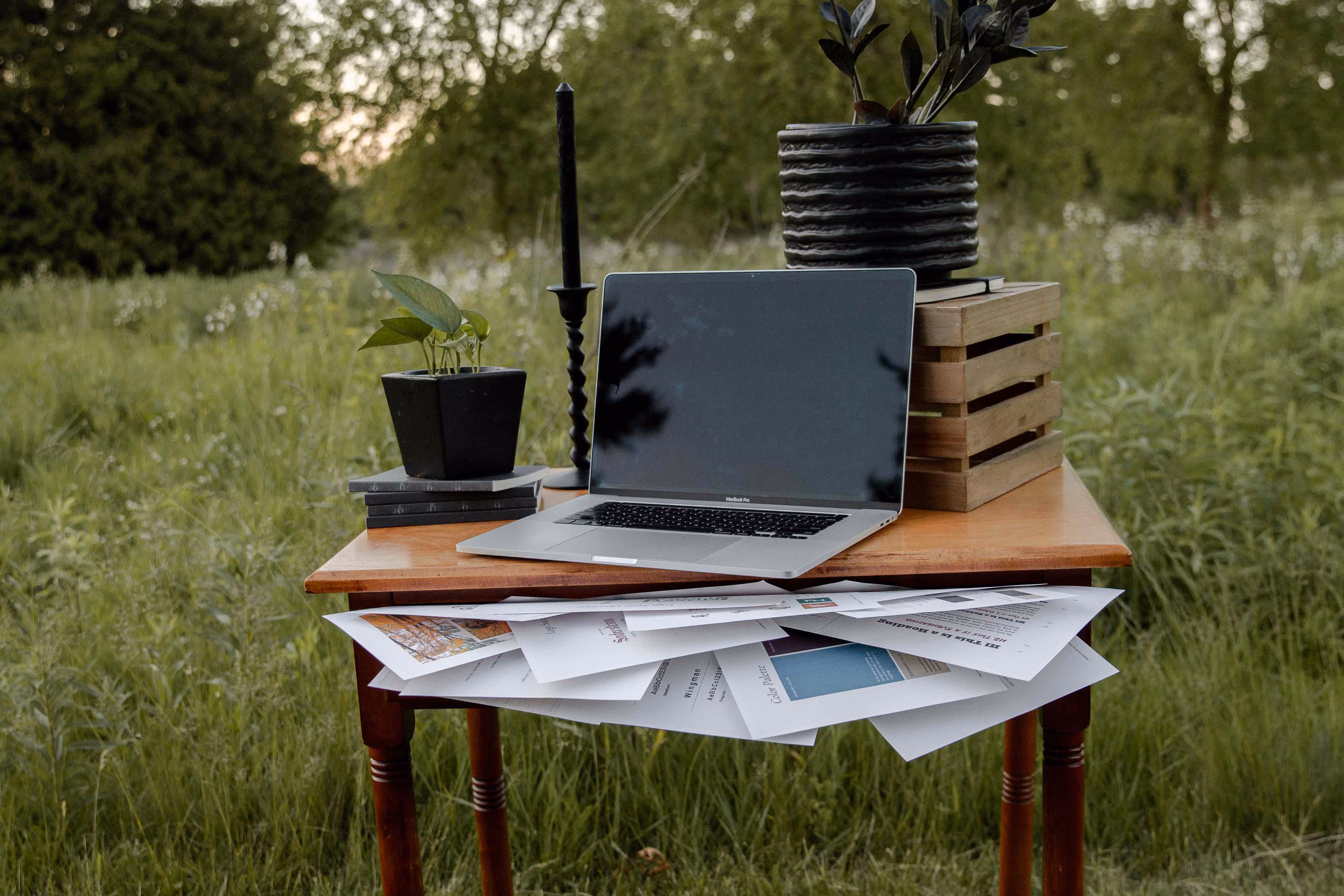 Outdoor wooden table with a laptop, scattered papers, two potted plants, and a black candle.