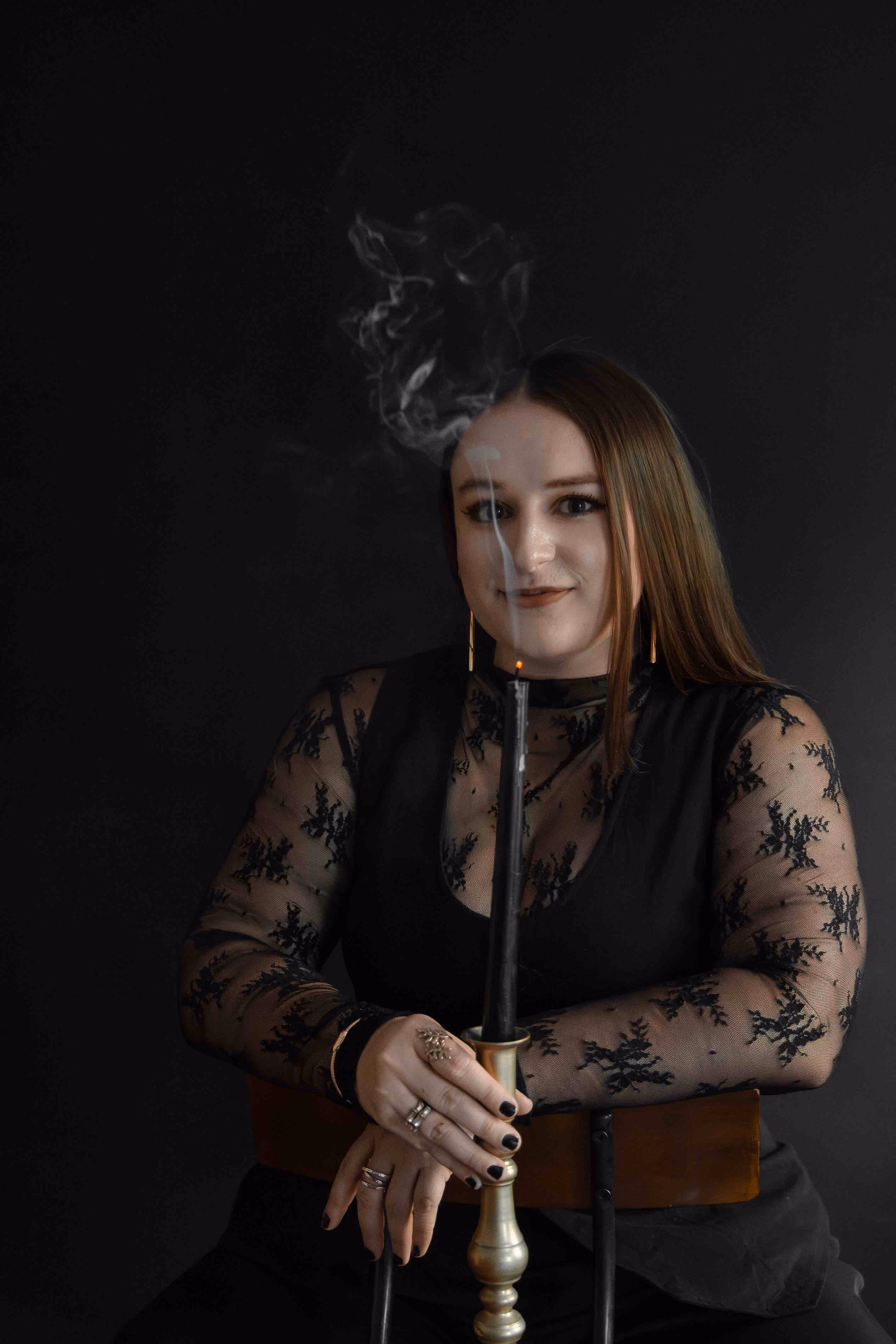 Woman in black lace top holding a candlestick with a smoldering black candle and smoke above.