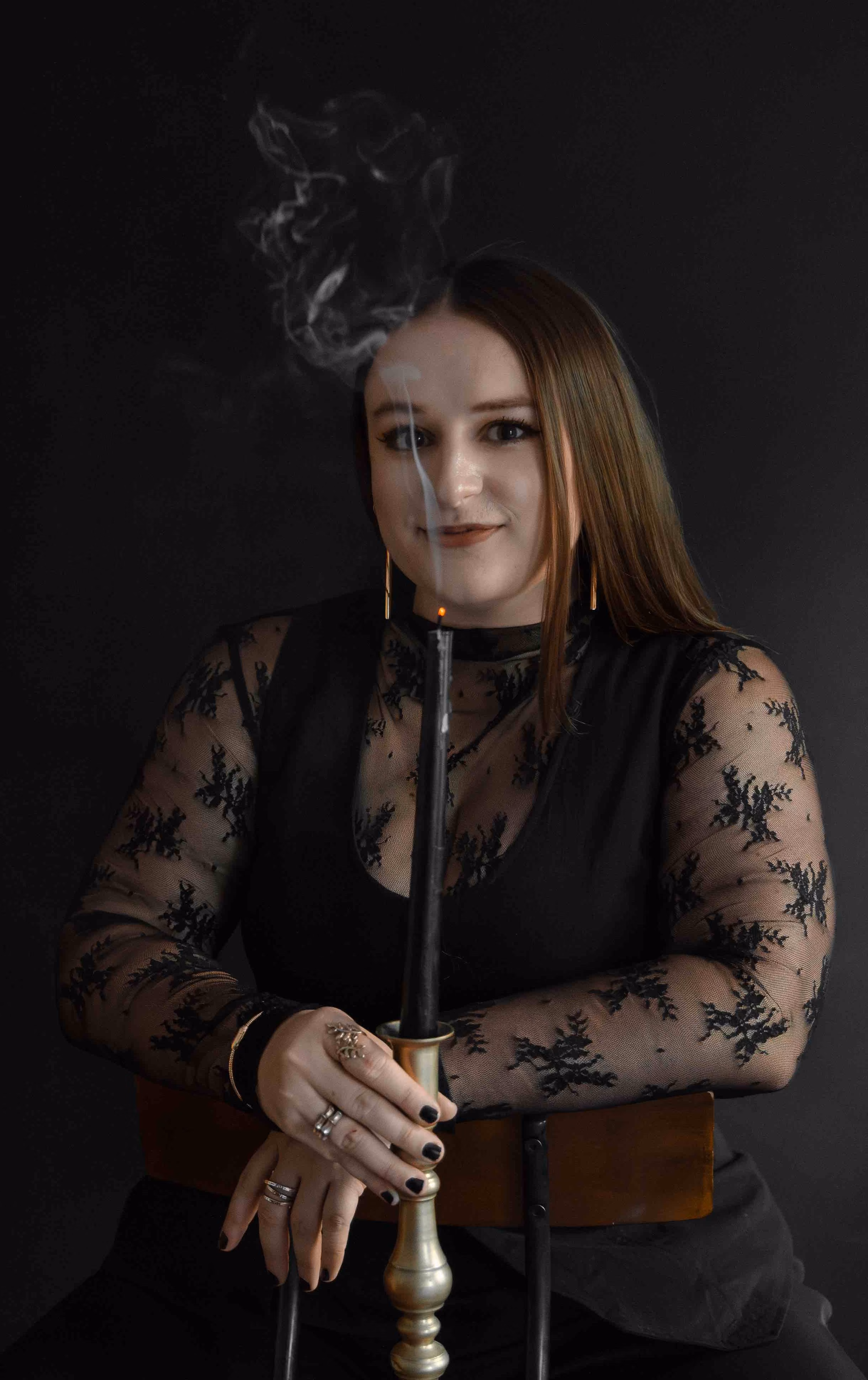 Woman in black lace top holding a smoking black candle in a brass candlestick against a dark background.