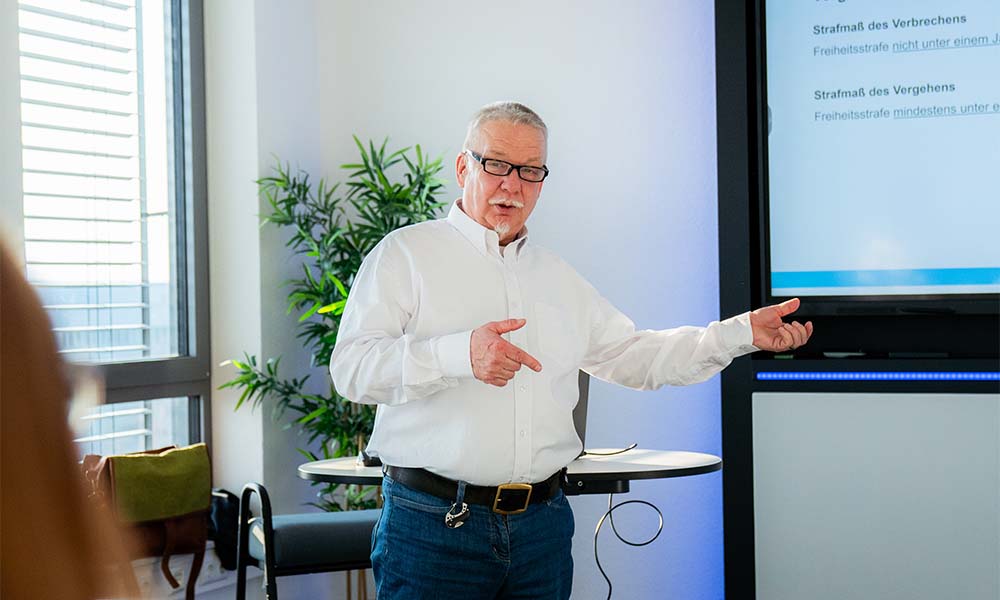 Älterer Mann in weißem Hemd und Brille hält Vortrag vor einem Präsentationsbildschirm in einem modernen Raum mit Pflanze und Fenster.