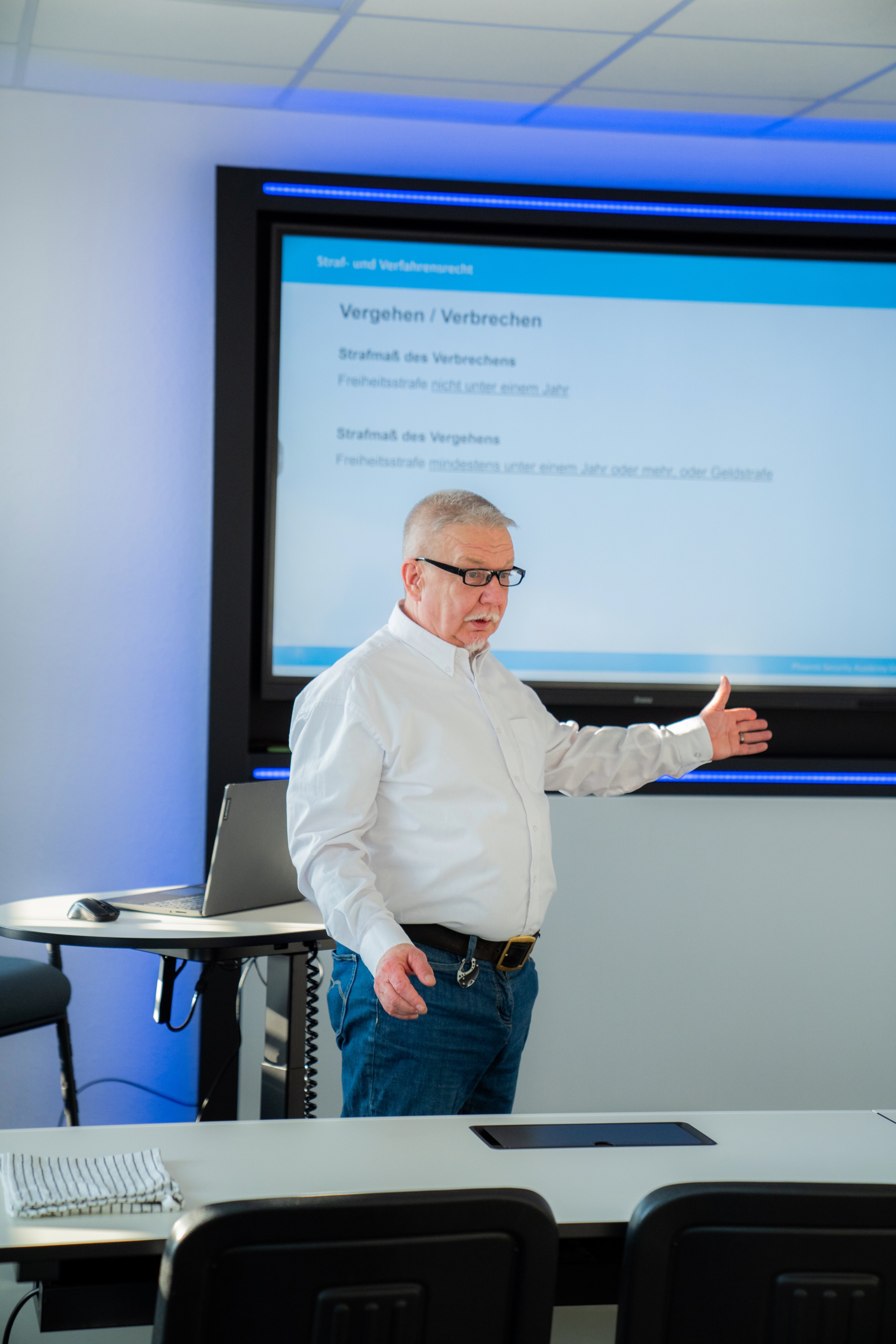 Älterer Mann mit Brille präsentiert vor einer Wand mit Text über Straftaten und Vergehen in einem Klassenzimmer.