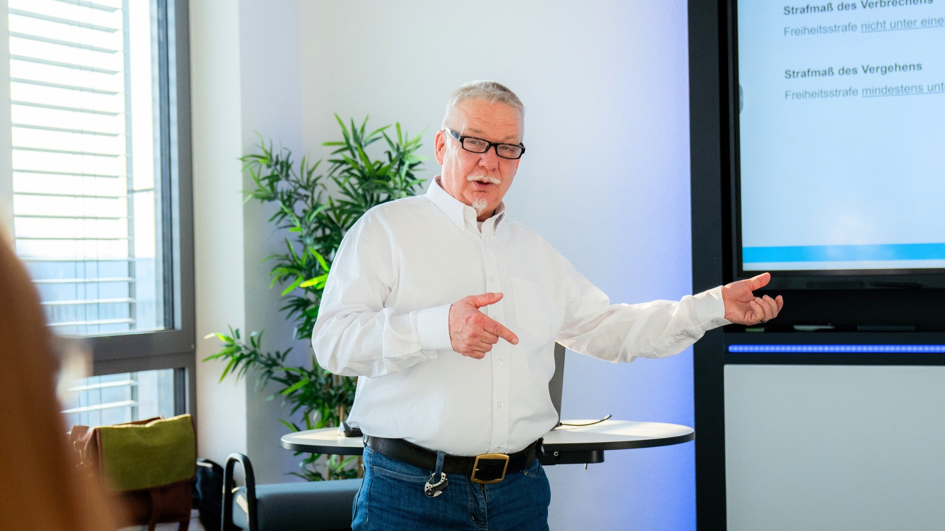 Älterer Mann mit Brille hält eine Präsentation vor Bildschirm mit Text in einem Büro.