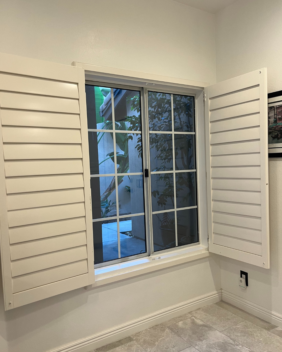 Before: Interior view of older sliding window with white grid panes and open white shutters