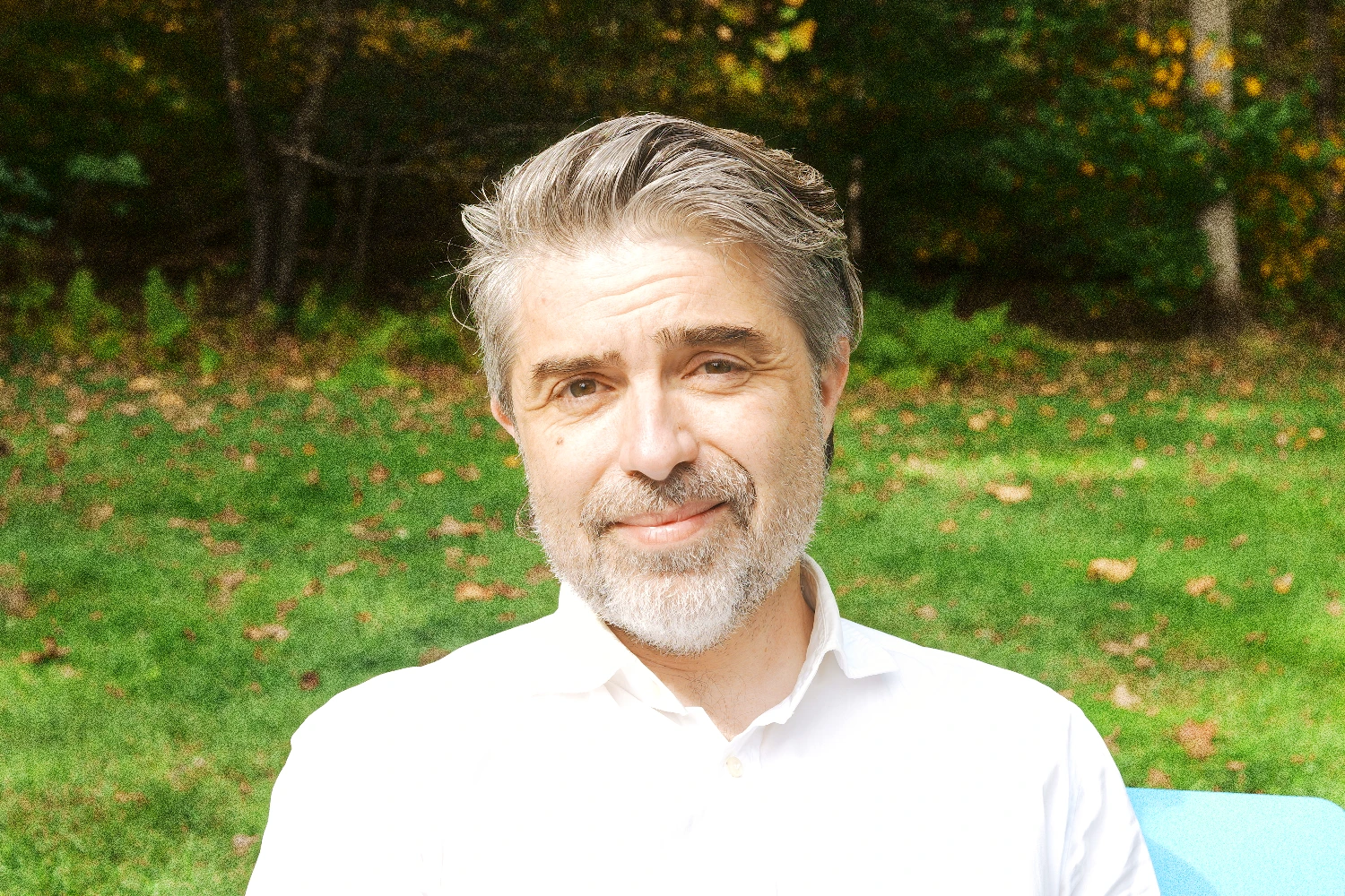 A photograph of a person seated outdoors in front of green grass and trees, wearing a white shirt, with their face covered by a large blurred rectangle.