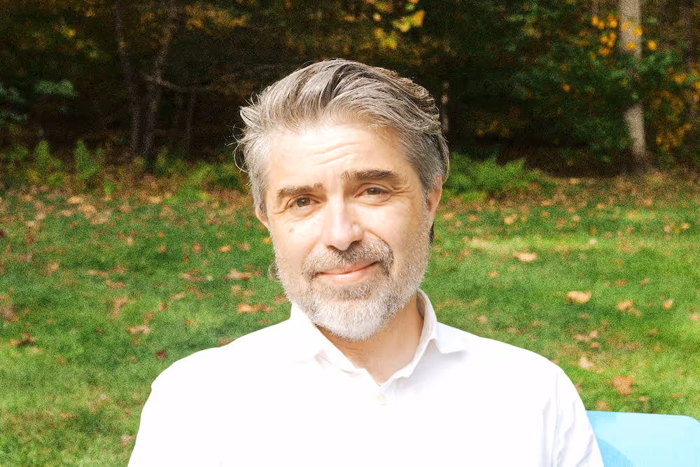 Middle-aged man with gray hair and beard smiling outdoors in front of green grass and trees.