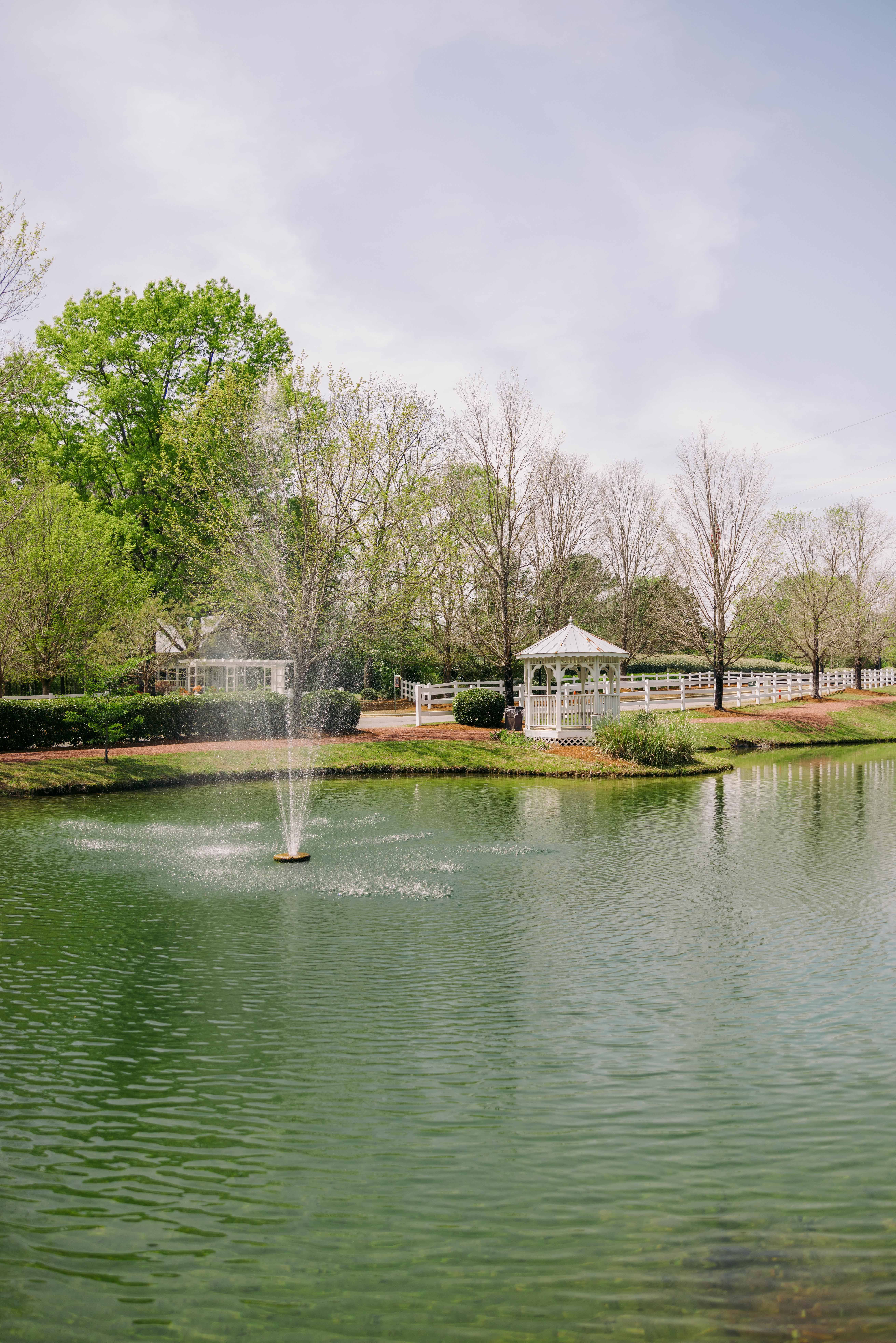 A serene pond with a central fountain, surrounded by lush trees and a white gazebo