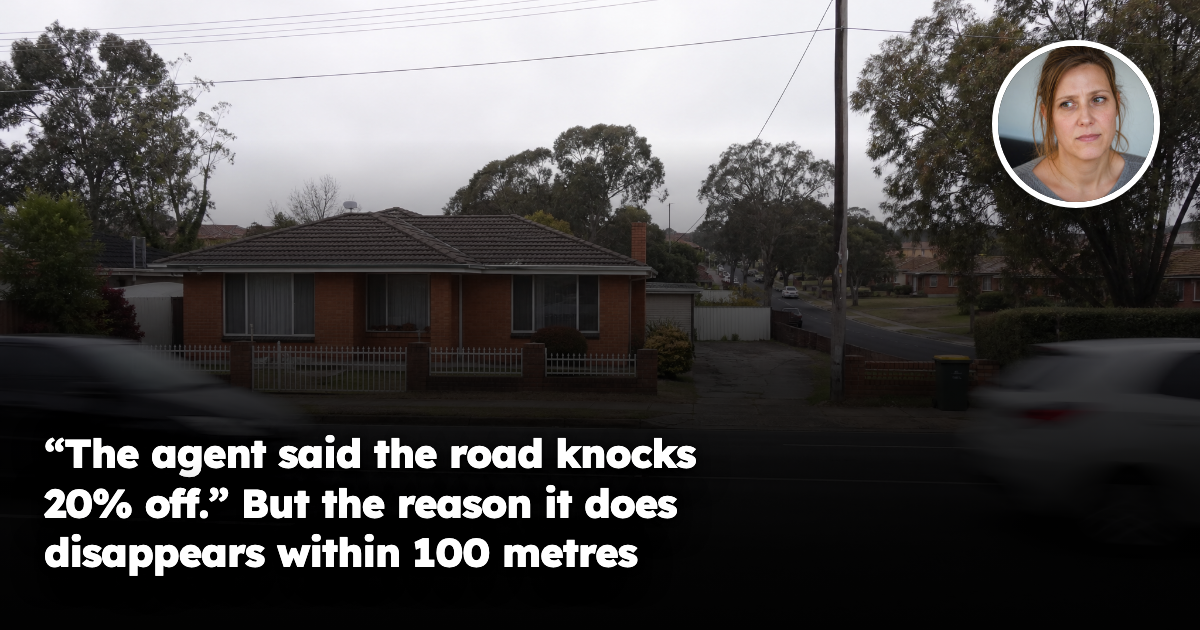 A modest single-storey suburban brick house sitting directly beside a busy four-lane arterial road, with cars blurred as they pass, a quieter leafy residential street visible further back under a flat overcast sky.