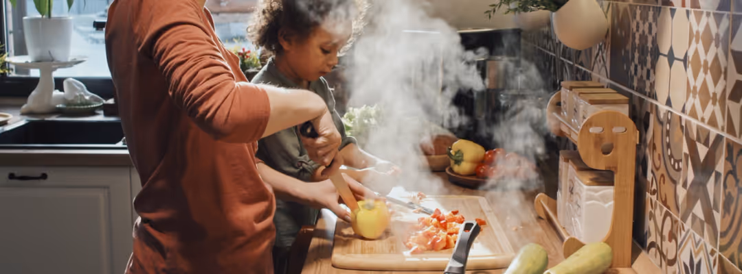Side view of an adult woman with her hair in a ponytail and a young child standing at a wooden countertop, slicing yellow bell peppers on a cutting board as steam rises from a sauté pan of mixed vegetables in a tiled kitchen.