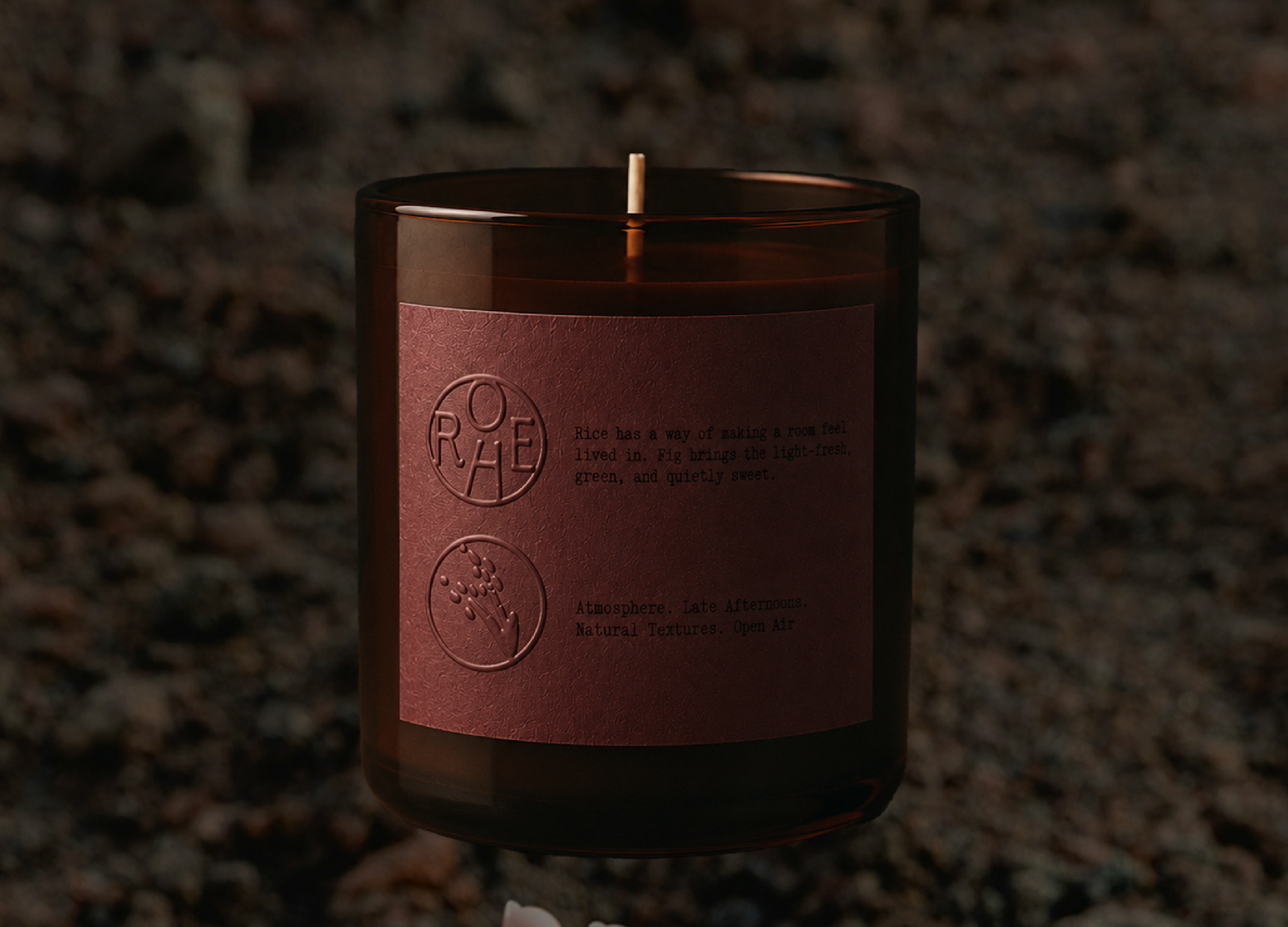 Brown glass candle with a purple label featuring text and circular embossed logos on a dark background.