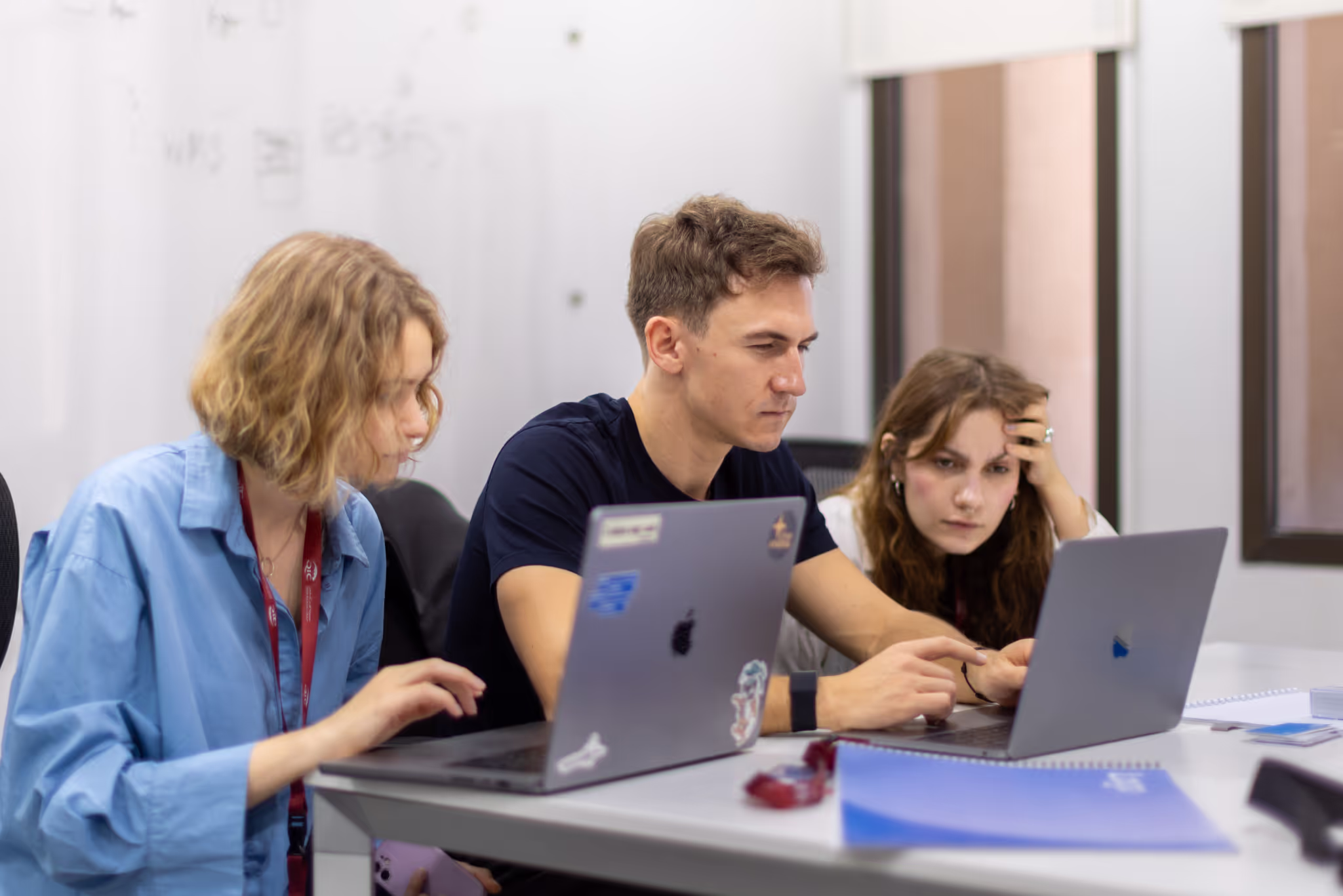 Three young adults working together on laptops at a white table in an office setting.