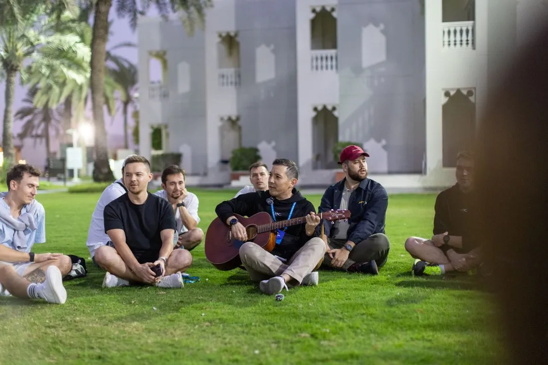 Group of young men sitting on grass in a park with one playing an acoustic guitar at dusk.
