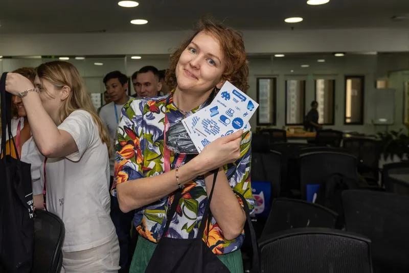 Smiling woman in a colorful floral jacket holding a brochure and a face mask in an office setting with people in the background.