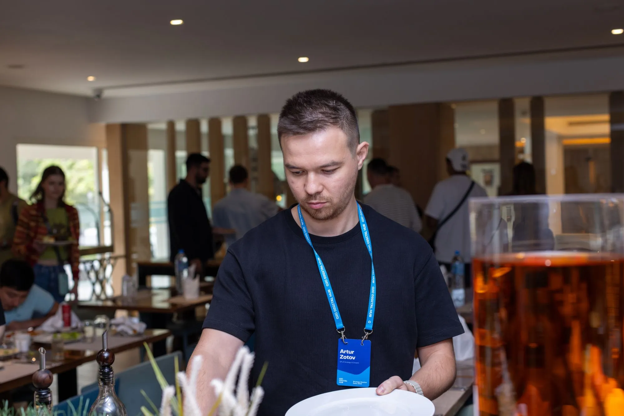 Man with short hair wearing a black shirt and blue conference badge holding a white plate indoors.