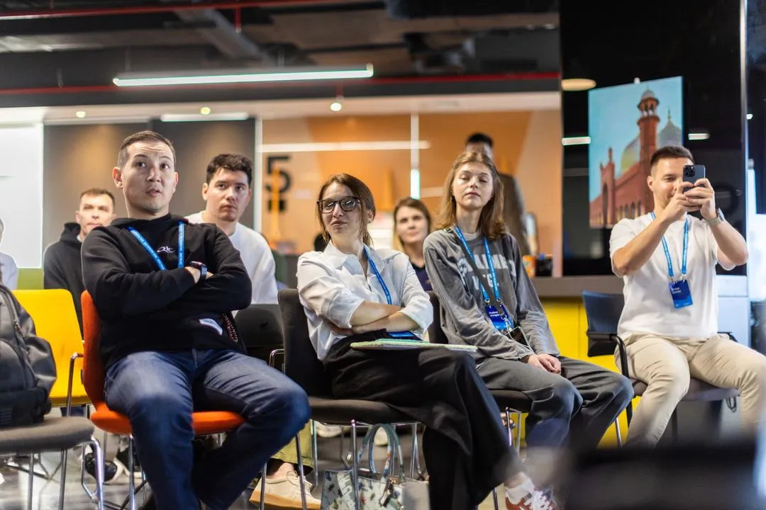 Group of young adults seated and attentively listening in a modern indoor setting, one person taking a photo with a smartphone.