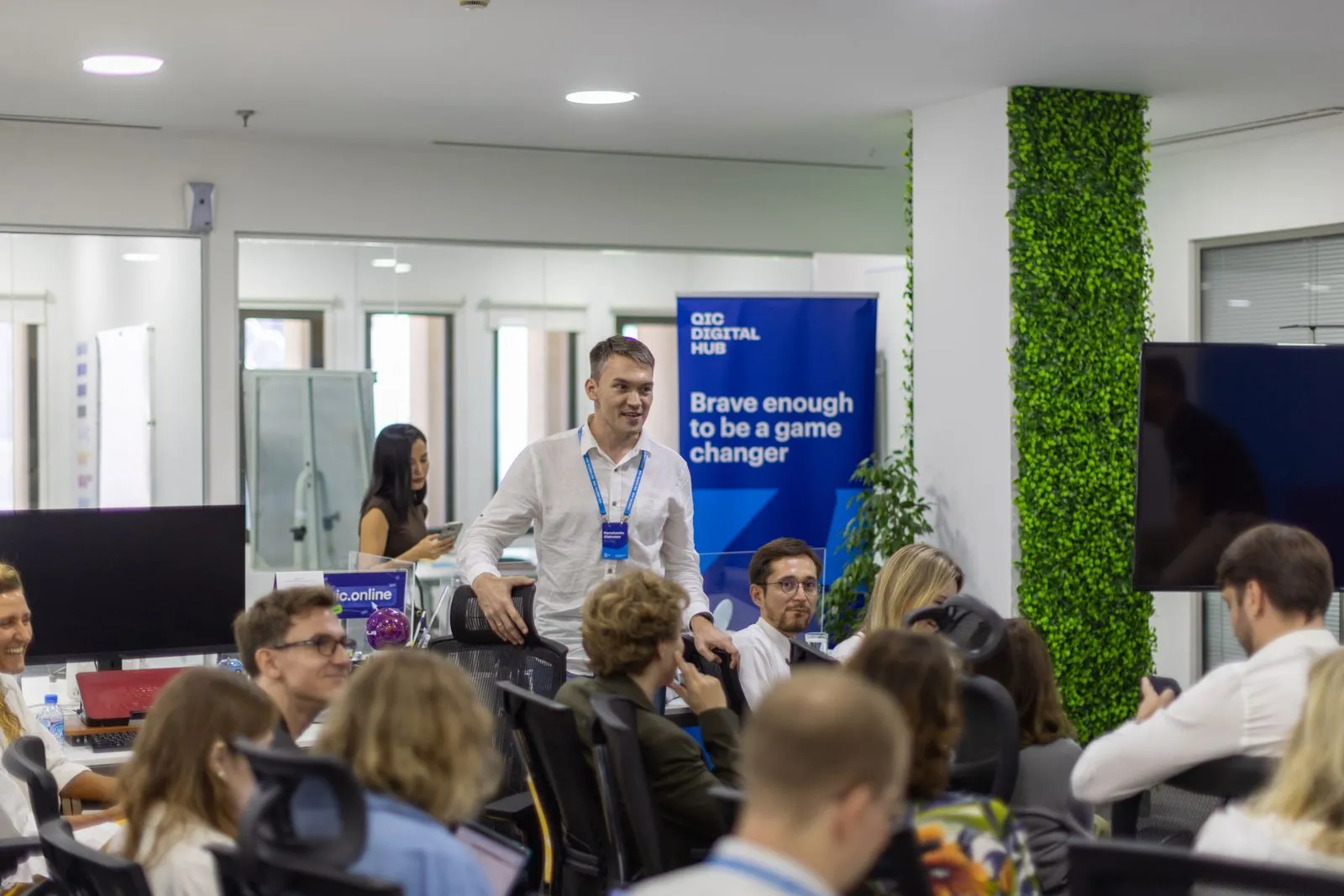 A man standing and speaking to a group of people seated in an office meeting room with a blue banner that reads 'Brave enough to be a game changer'.