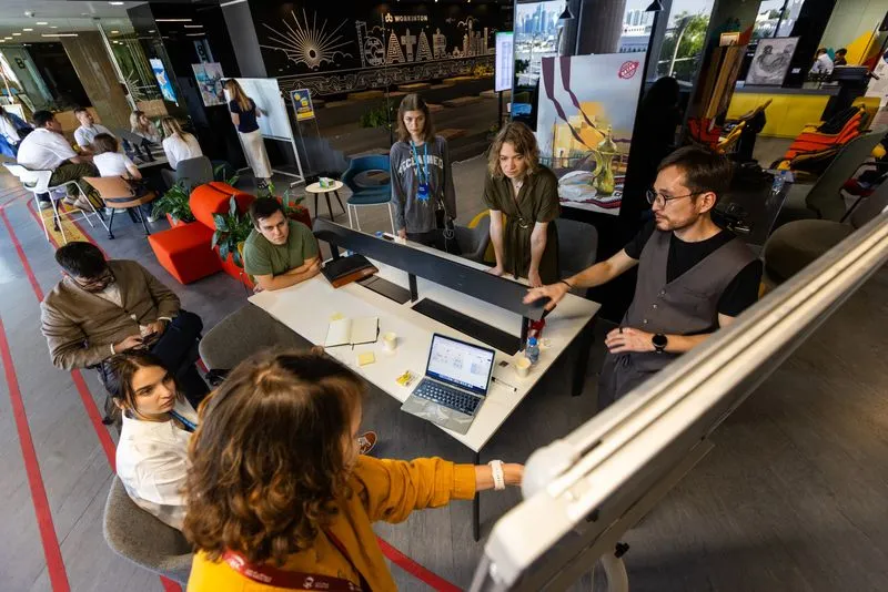 Group of six people engaged in a discussion around a table with laptops and notebooks in an open office space.