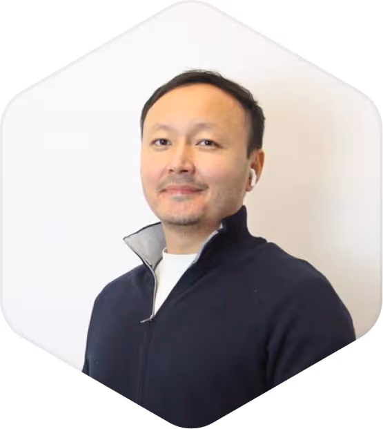 Man with short dark hair wearing a dark zip-up sweater and white earbuds, smiling slightly against a white background.