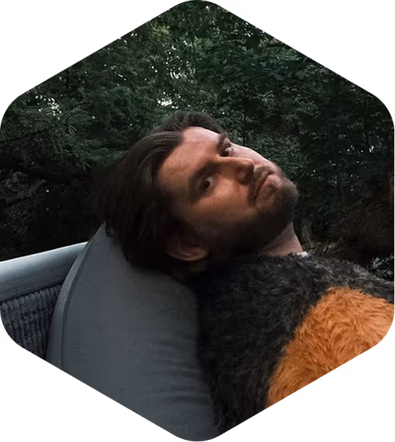 Man with dark hair and beard reclining on a chair outdoors with trees in the background.