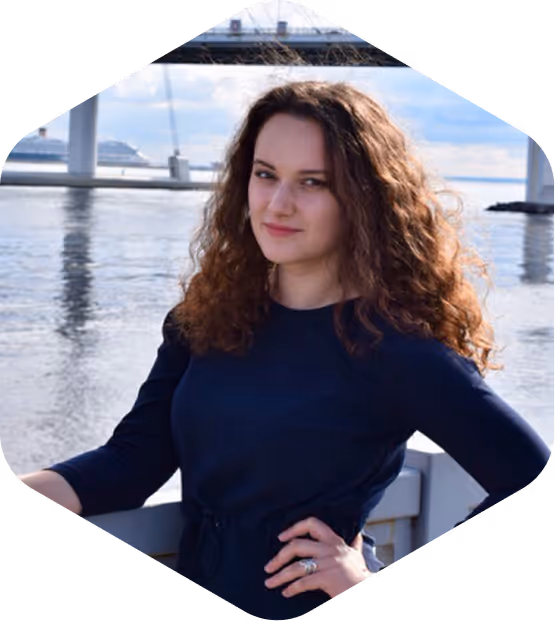 Young woman with curly brown hair and a navy blue dress standing by a waterfront with a bridge in the background.