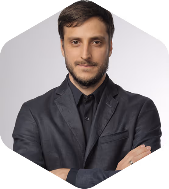 Man with short brown hair and beard, wearing a black shirt and blazer, standing with arms crossed against a gray background.