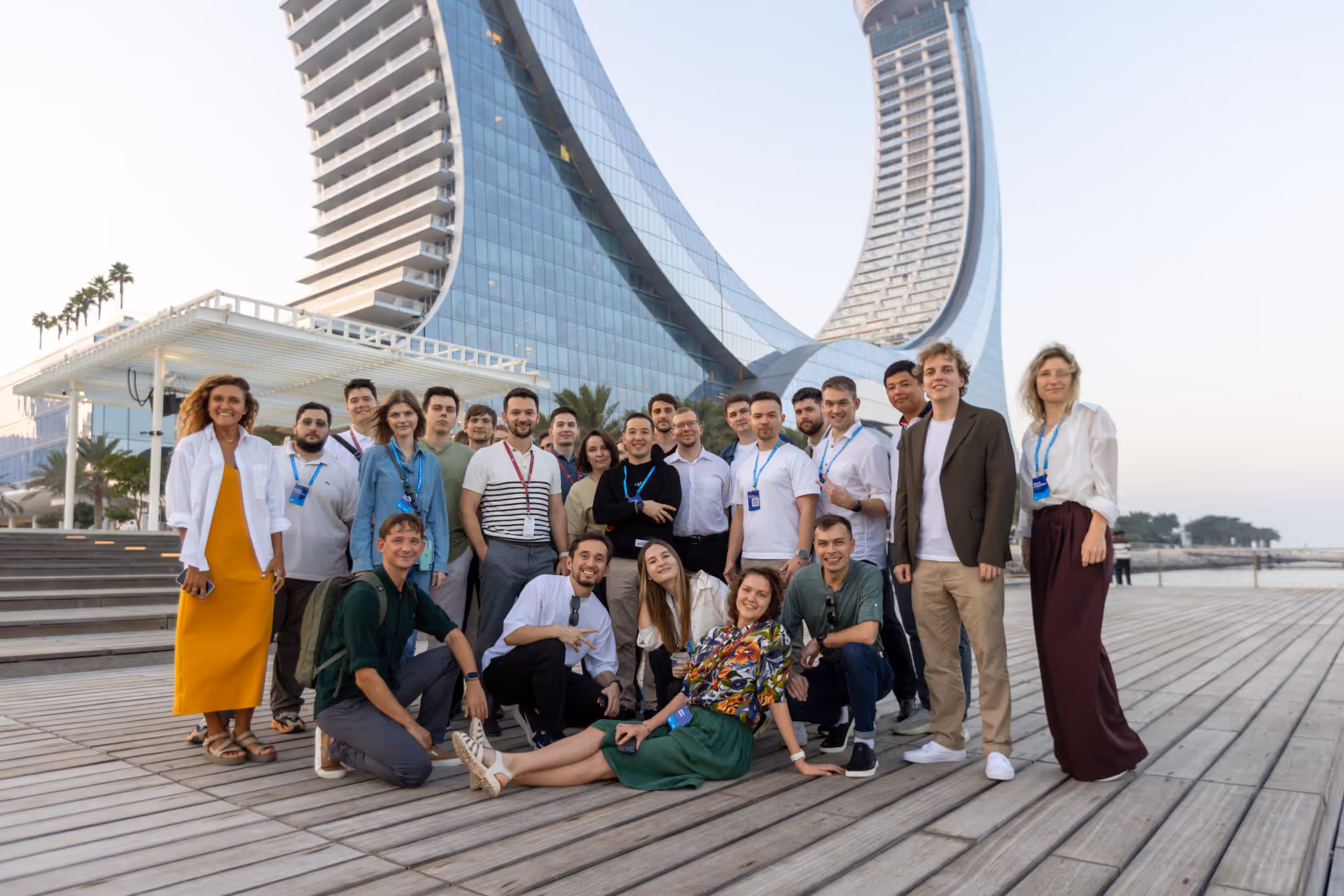 Group of diverse young adults posing together on a wooden deck with modern curved glass buildings in the background during daylight.