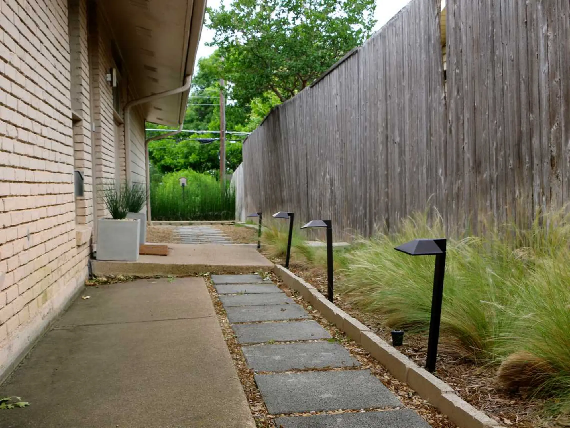 A serene walkway lined with grass and plants, leading up to a textured brick wall in the background.
