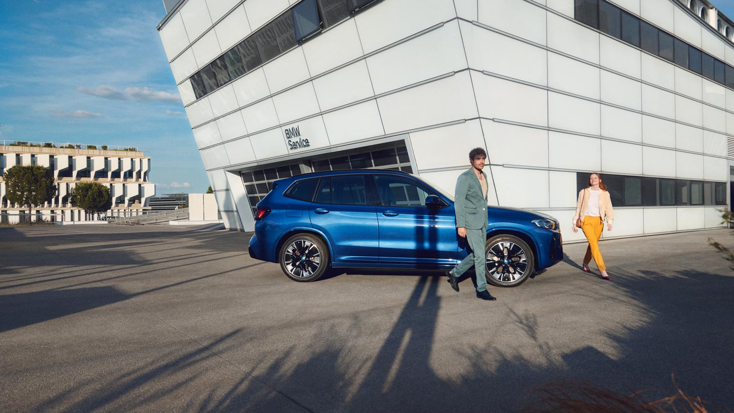 Blauer BMW iX3 vor einem futuristischen Gebäude mit einem Mann und einer Frau– Modernität und Technologie im Autohaus BMW Köhler in Karlstadt