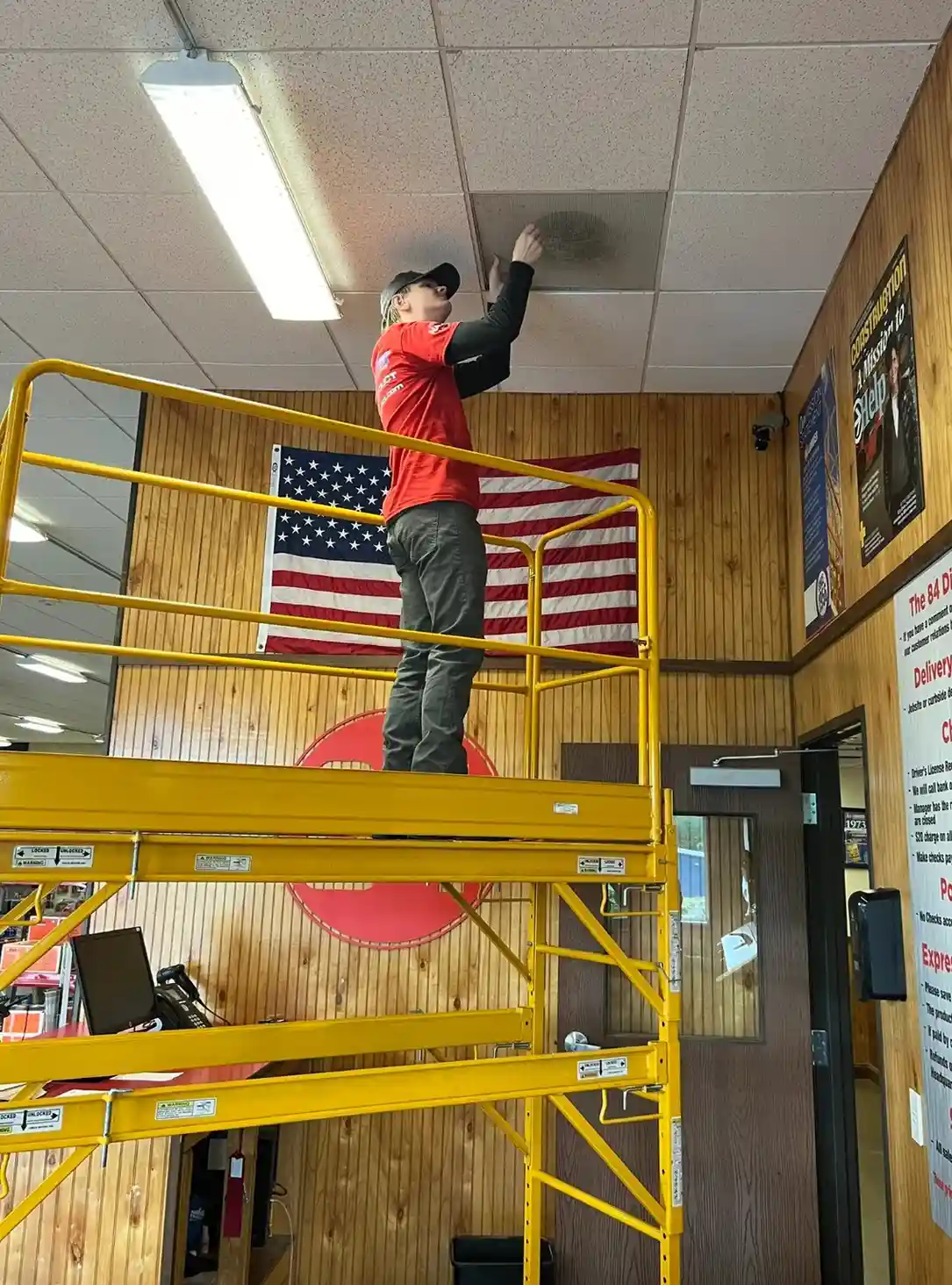 A technician, possibly identified as Jakob, is standing on yellow scaffolding and reaching up to service a ceiling-mounted unit or duct, with an American flag visible in the background.