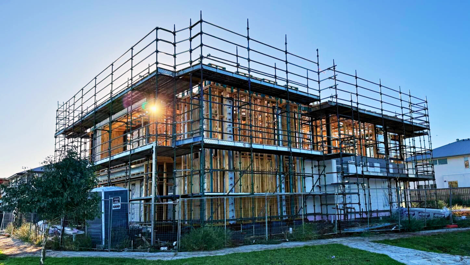 A construction worker wearing a yellow hard hat and a reflective safety vest is standing on a wooden frame of a building