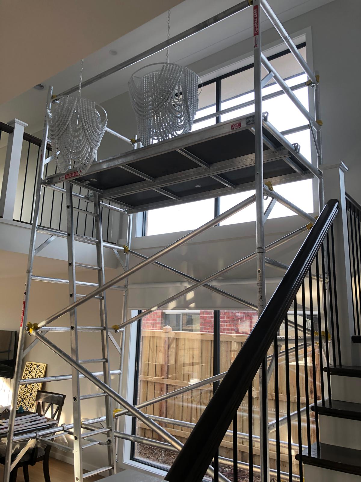 Interior view of a living area with mobile scaffolding set up near a large window and two white beaded chandeliers hanging above.