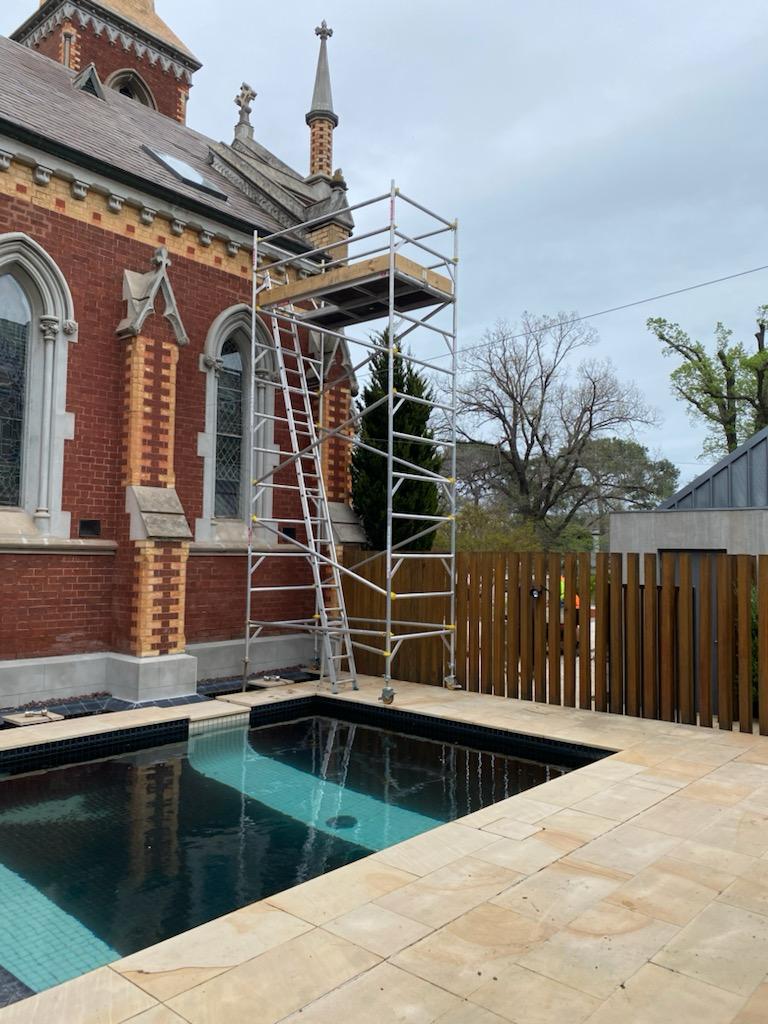 Mobile  scaffolding with a ladder set up beside a red brick building near a tiled pool under a cloudy sky.