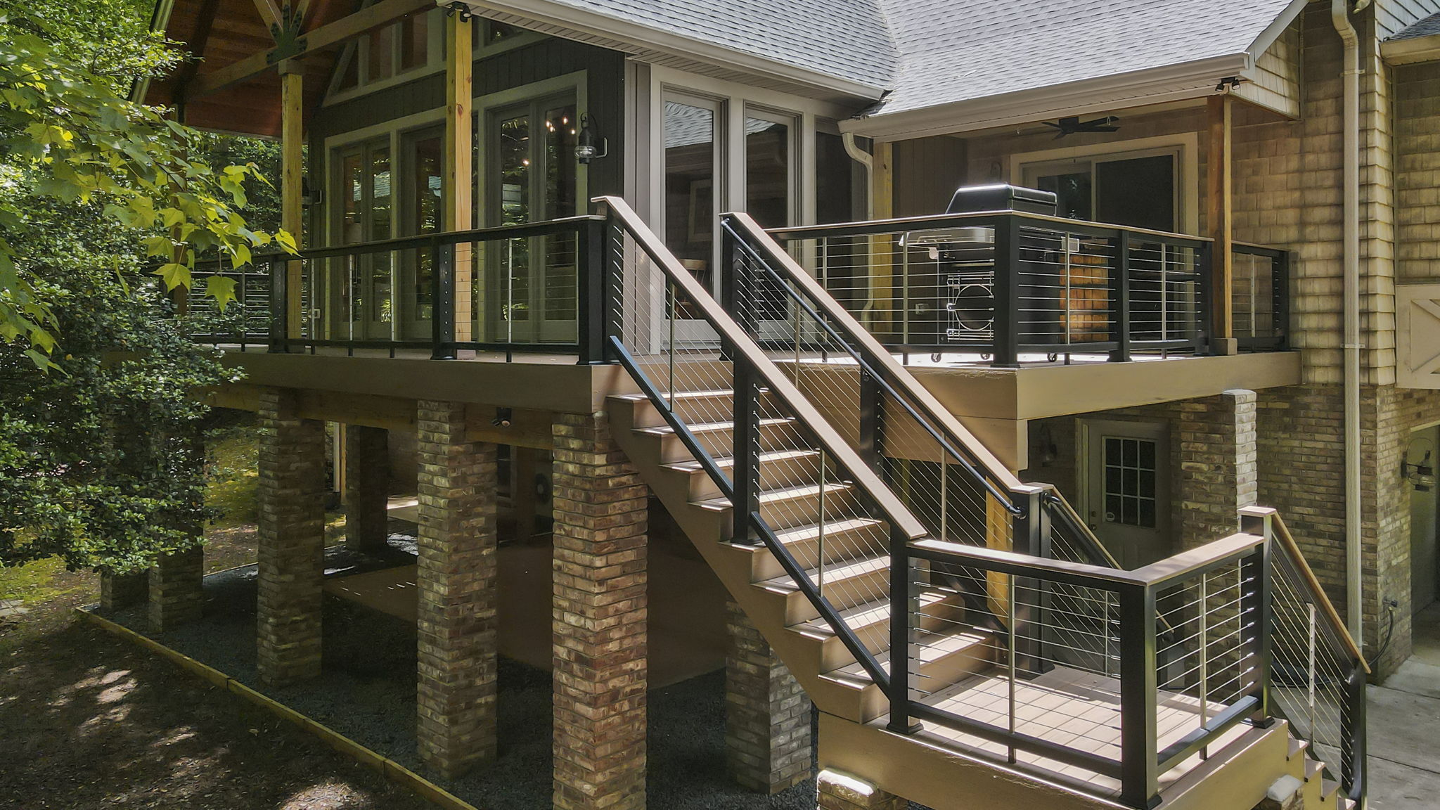 Elevated wooden deck with black metal railing and staircase attached to a house with brick pillars and many windows.