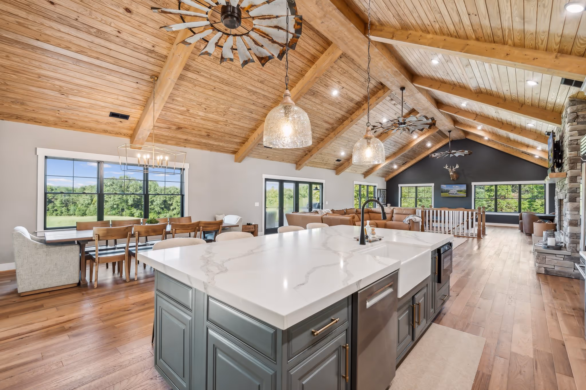 Open kitchen and living area with wooden vaulted ceiling, large island with marble countertop, dining table, and large windows showing greenery outside.