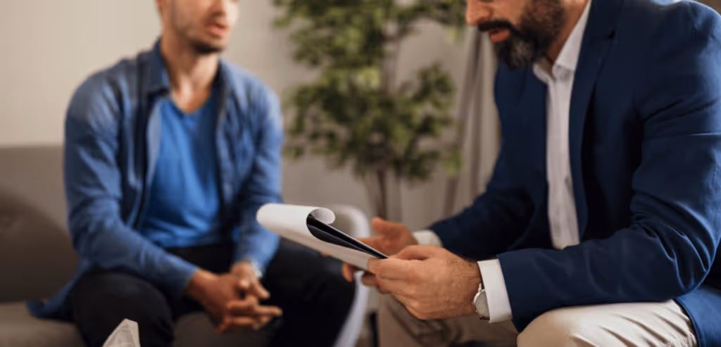 A man sits on a couch with his hands clasped. Another man sits in front of him looking down at a journal