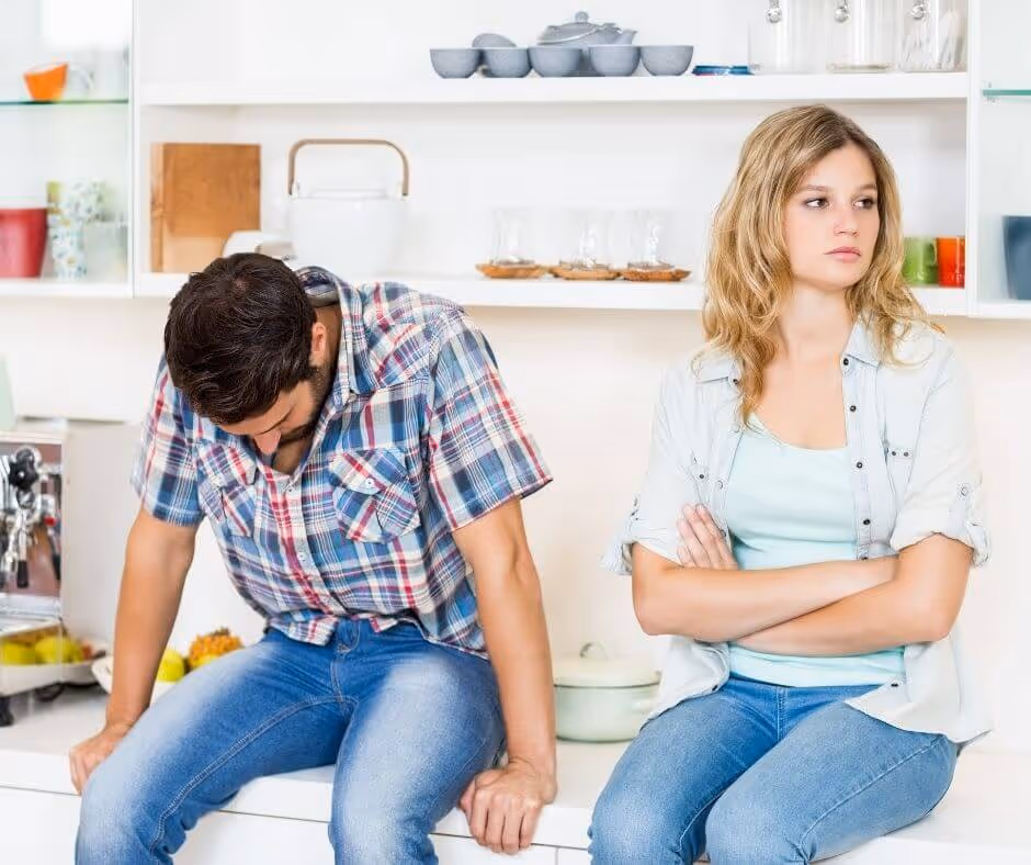 A couple sitting on the kitchen counter. The man is looking down and the woman is folding her arms looking away