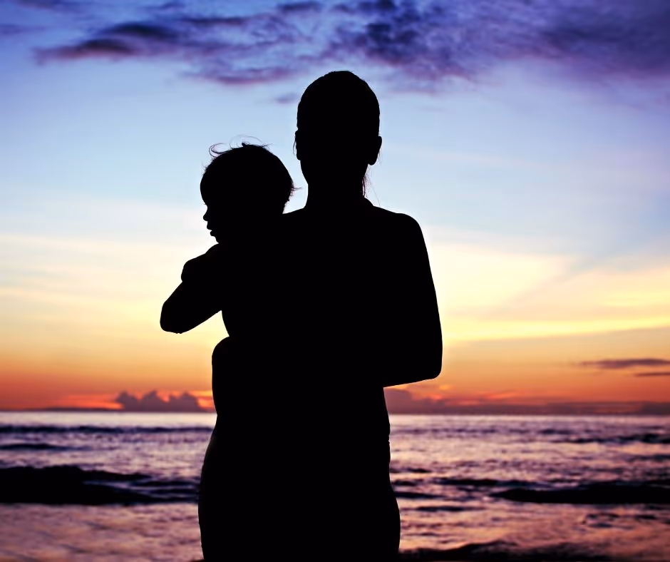 A parent holds their infant looking out over the ocean at sunsent